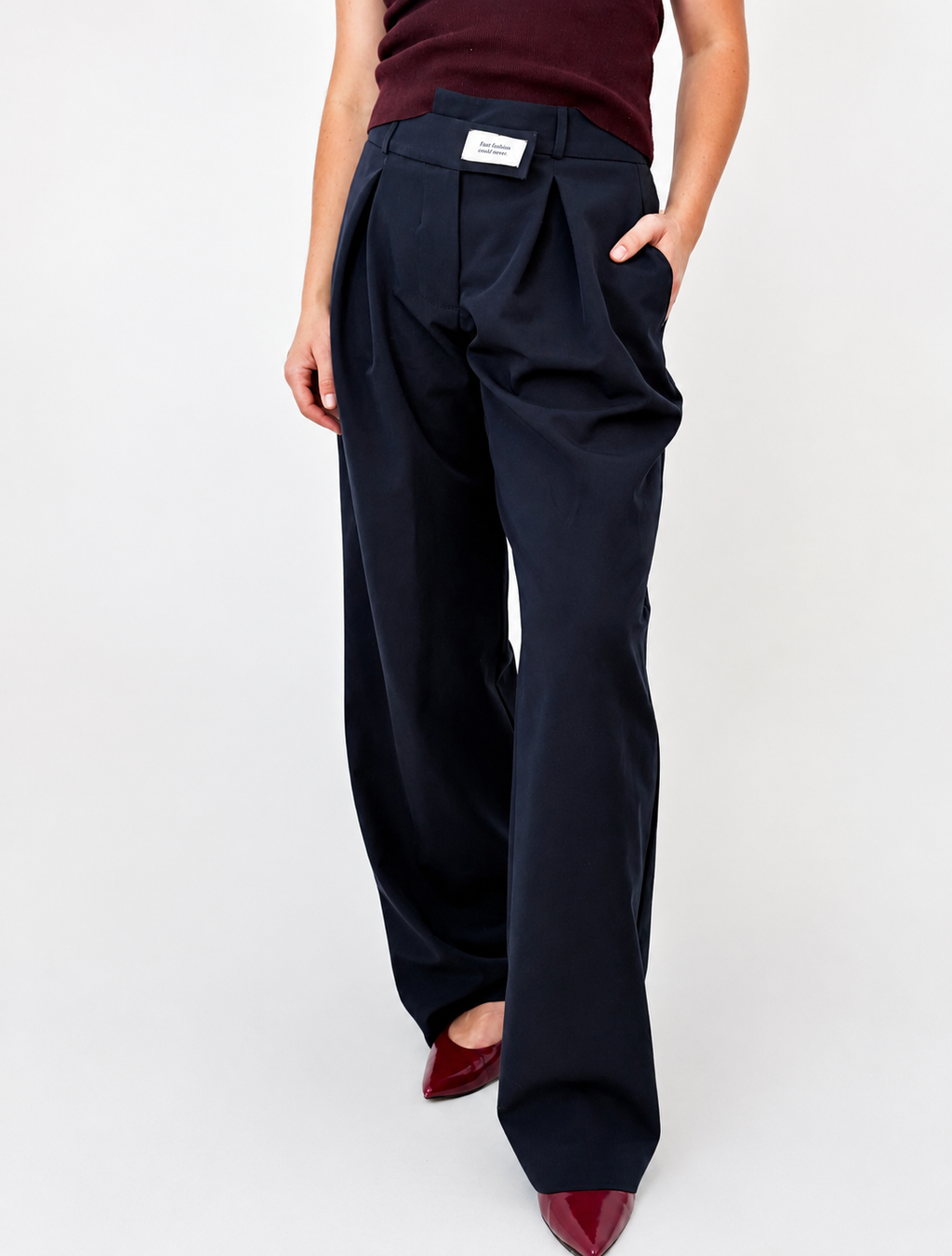 Close-up of the Founder's Cut trousers in navy, featuring a wide-leg silhouette, front pleats, side pockets, and the signature asymmetric waistband detail with the Courthill XIX label. Styled with a burgundy knit top and pointed burgundy heels.
