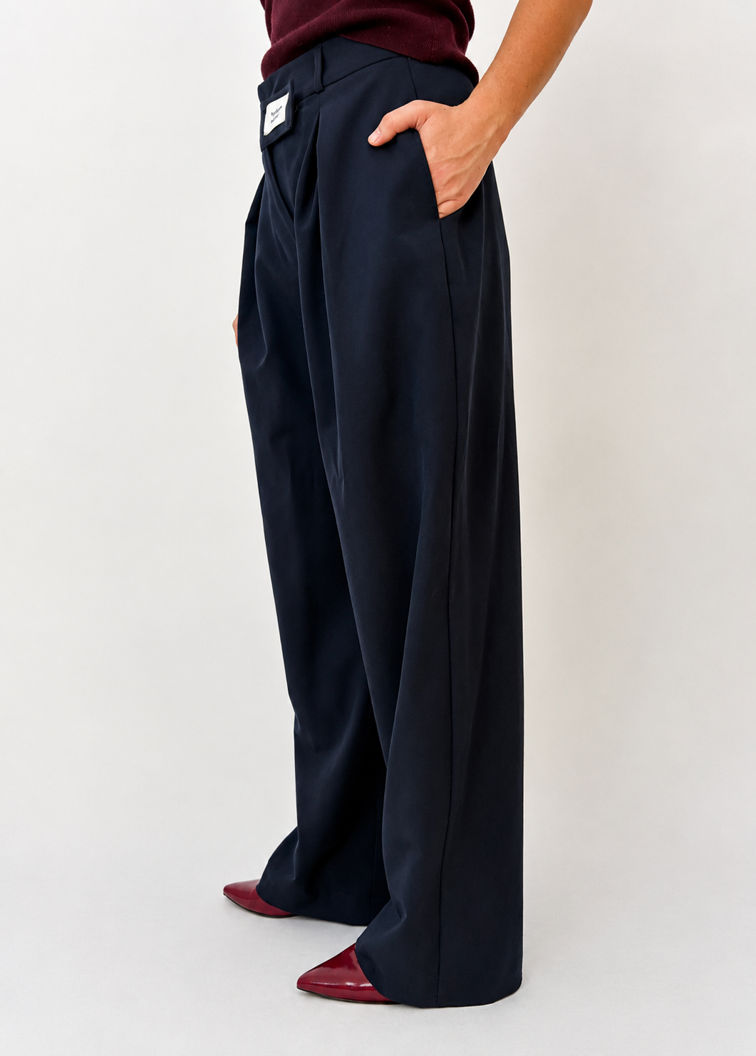 Side view of the Founder's Cut trousers in navy, showcasing the relaxed wide-leg fit, deep front pleats, side pocket, and signature asymmetric waistband detail. Styled with a burgundy knit top and pointed burgundy heels.