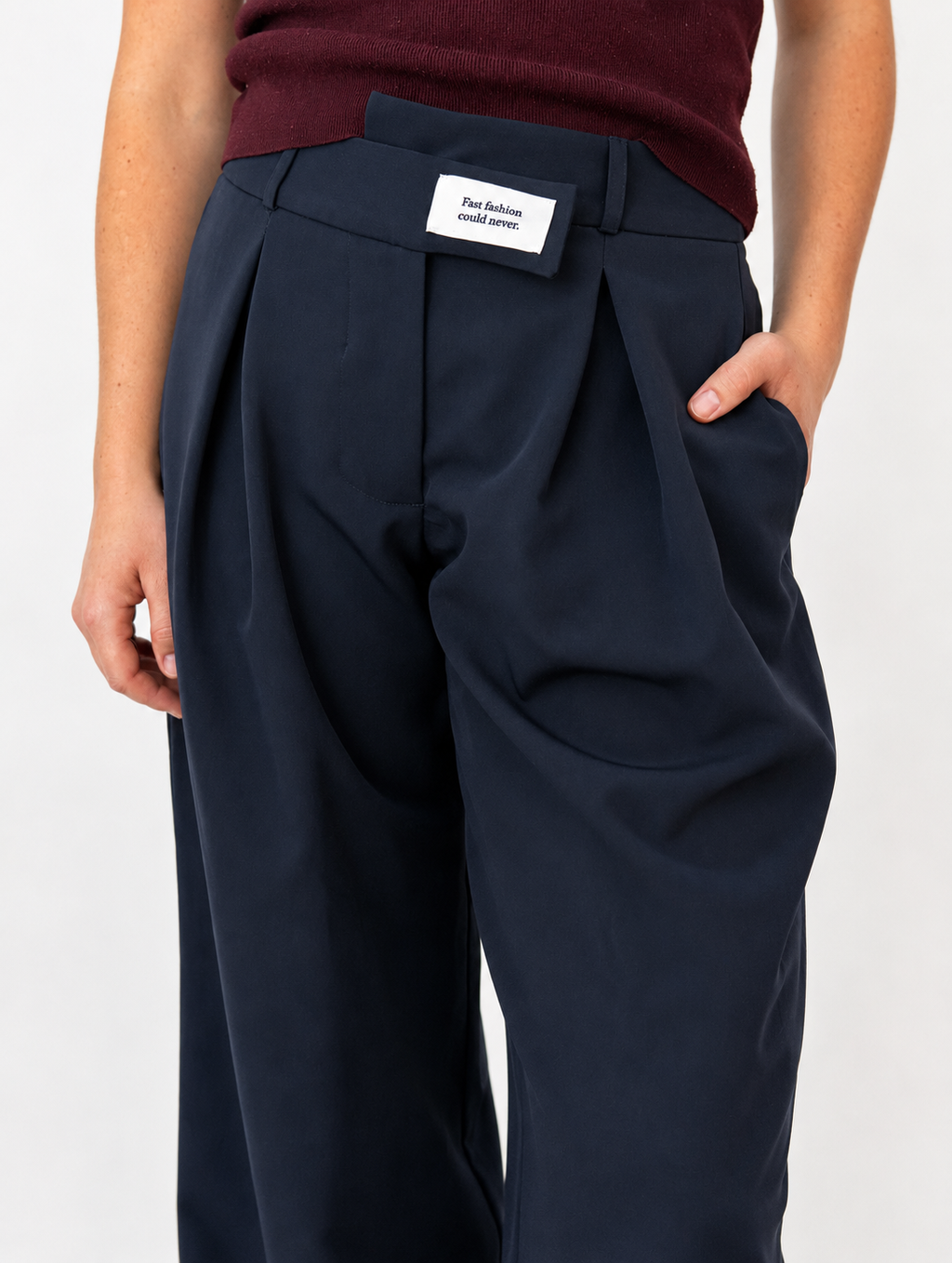 Close-up of the Founder's Cut trousers in navy, highlighting the signature asymmetric waistband with woven statement label, tailored front pleats, and side pocket. Styled with a burgundy knit top.