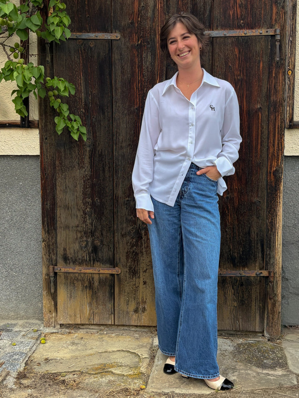 Full-length outdoor shot of the Essence Shirt styled with relaxed wide-leg blue jeans. The embroidered CH logo on the chest, oversized silhouette, and fluid fabric create a timeless, effortless look, framed by a rustic wooden door.