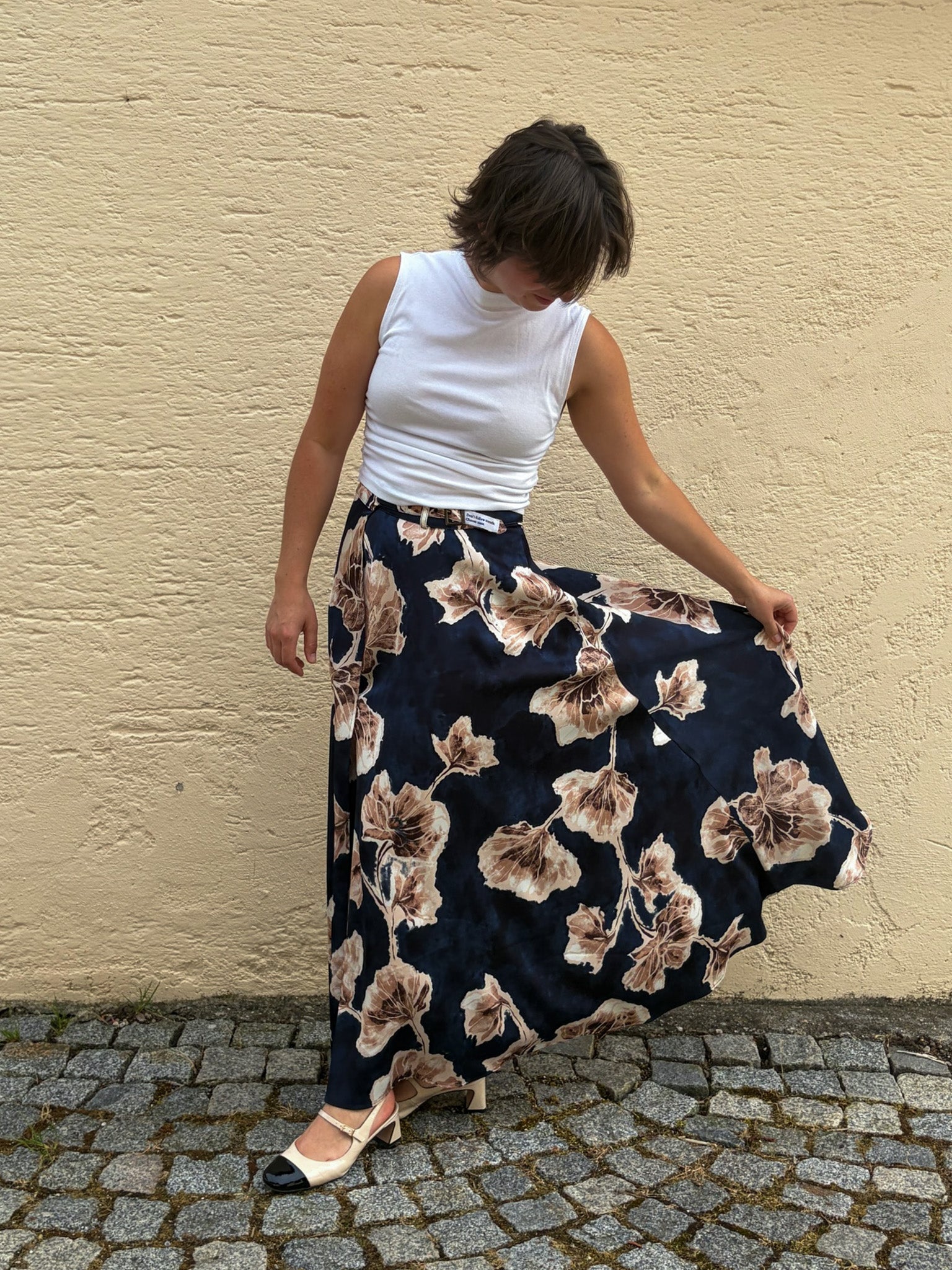 Model trägt den Spring Flow Maxirock mit floralem Print und einem weißen ärmellosen Oberteil. Sie hält den Rock leicht zur Seite, wodurch der fließende Fall des knitterarmen Travel-Fabrics und die großzügige Weite des Designs besonders gut zur Geltung kommen.