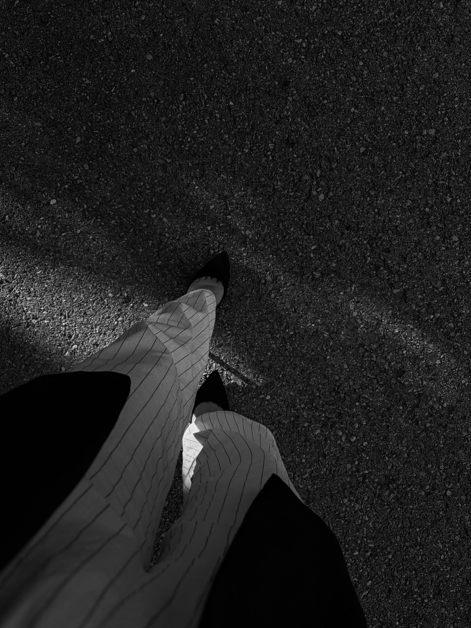 Schwarz-weißes Foto aus der Ich-Perspektive einer gehenden Person mit einer weißen, fein gestreiften Hose und spitzen Schuhen auf Asphalt. Der Fokus liegt auf dem minimalistischen Outfit und der Bewegung im Alltag.