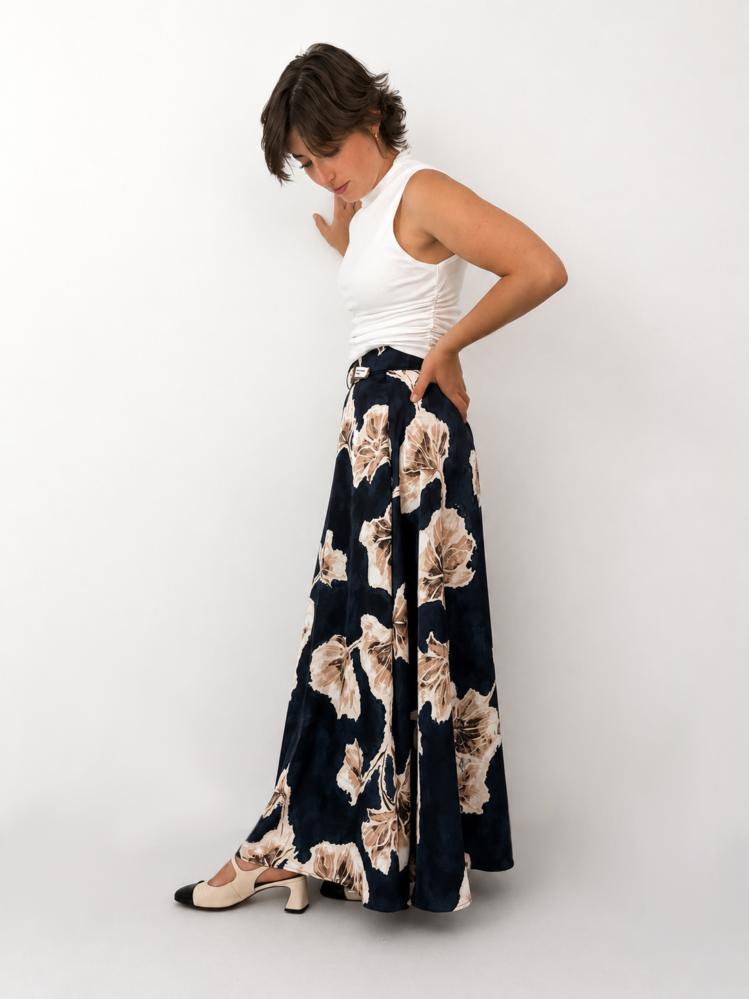 Model trägt den Spring Flow Maxirock mit floralem Print in Kombination mit einem weißen ärmellosen Oberteil. Die Seitenansicht zeigt den fließenden Fall des Rocks, den individuell verstellbaren Bund und die elegante Silhouette vor einem schlichten weißen Hintergrund.