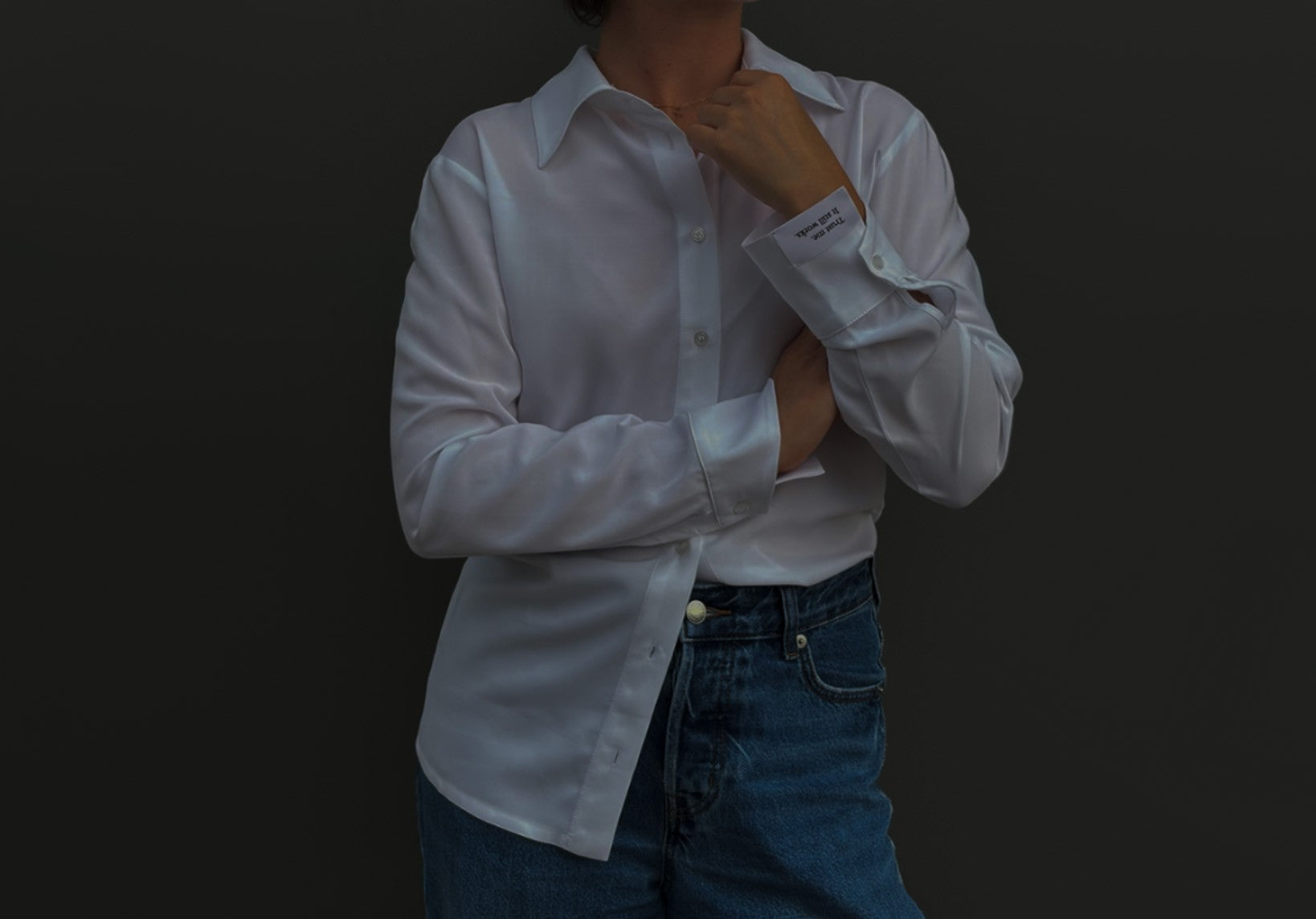 Weiße Damenbluse mit klassischem Kragen und langen Ärmeln, kombiniert mit einer blauen Jeans. Die Person hält eine Hand am Kragen, wodurch die breite Manschette mit eingearbeitetem Schriftzug sichtbar wird. Dunkler Hintergrund mit Fokus auf den Stoff, die Details der Bluse und die Manschette.