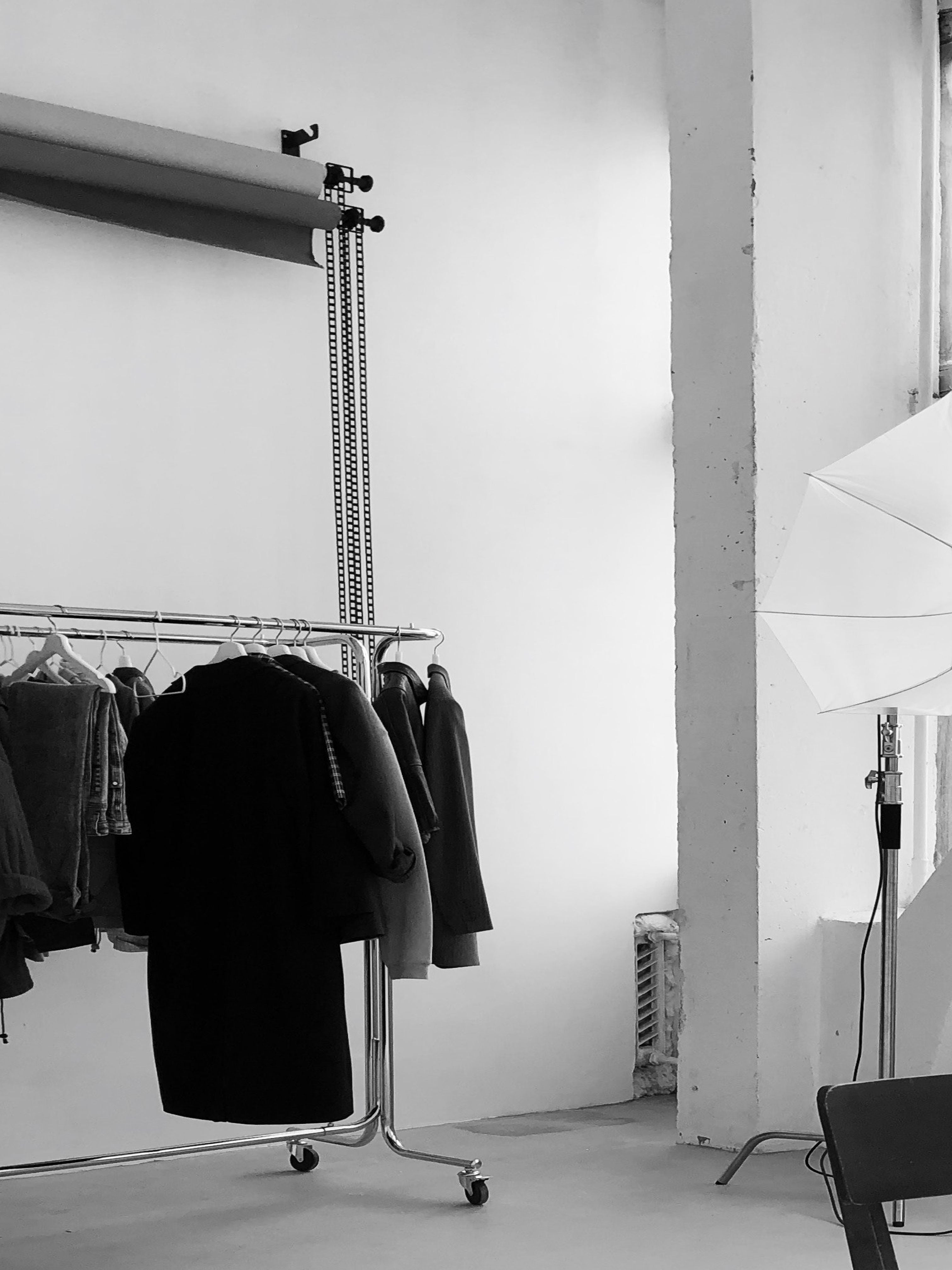 Schwarz-weißes Bild eines Fotostudios mit einer Kleiderstange voller Kleidungsstücke, darunter Mäntel, Blazer und Hosen. Im Hintergrund befinden sich eine Papierhintergrundrolle und Studiobeleuchtung. Die minimalistische Szene vermittelt den Entstehungsprozess einer Modekollektion und den Blick hinter die Kulissen von Courthill XIX.
