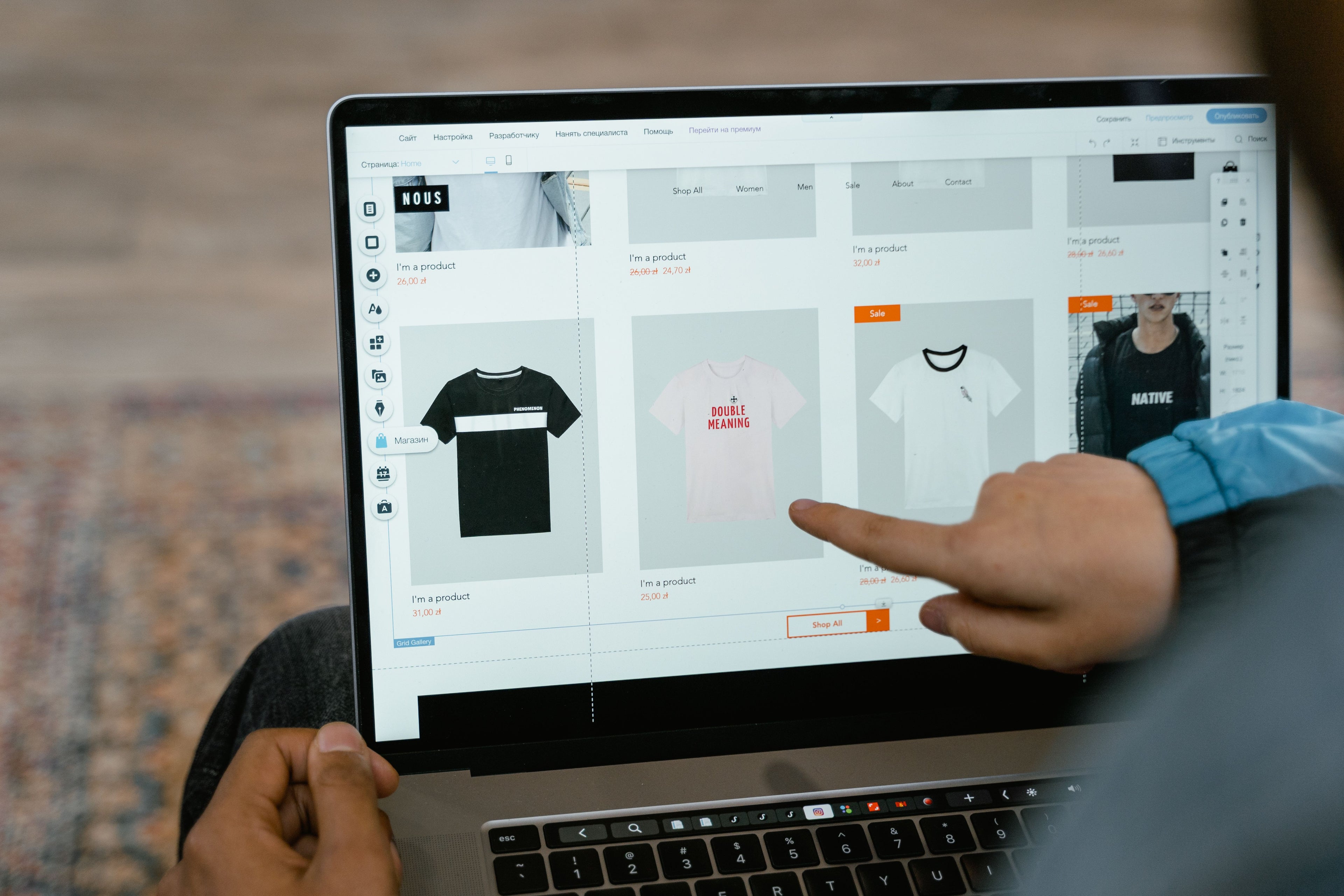 Nahaufnahme eines Laptops während der Gestaltung eines Onlineshops. Eine Person zeigt auf die Produktübersicht mit verschiedenen Kleidungsstücken und bearbeitet das Shop-Layout. Das Bild steht für die Entwicklung und den Aufbau des Courthill XIX Onlineshops.